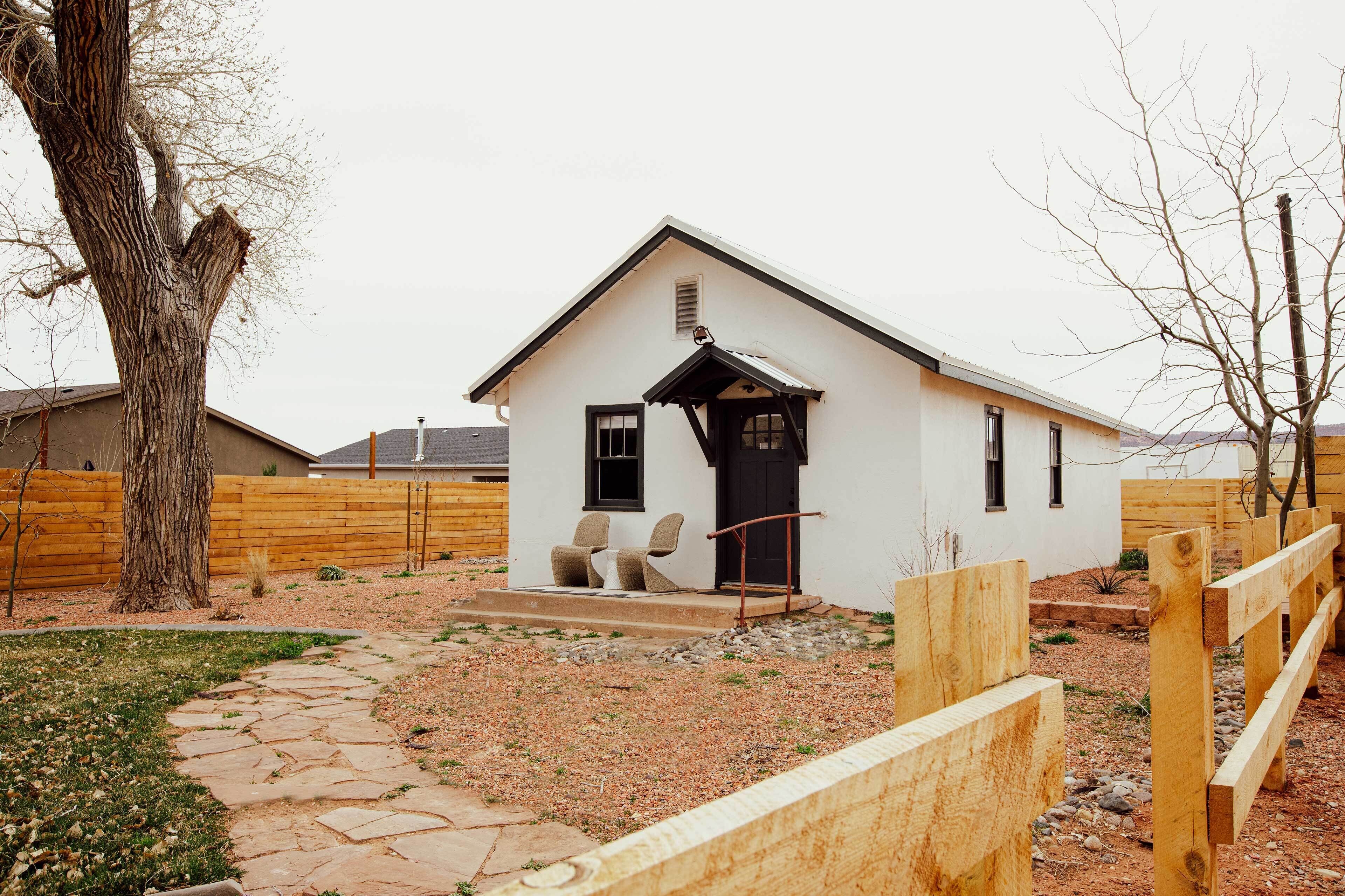 A small white building with a black door and windows, a stone pathway leading to the entrance, and a wooden fence surrounding the property.
