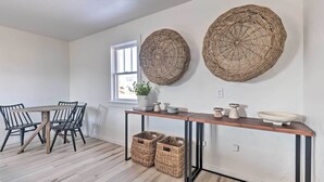 A dining area with a wooden table and chairs, a woven wall hanging, and wicker baskets under the table. - Modern Kanab Cottage w/ View < 1 Mi to Hiking! (Kanab)