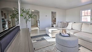 A modern living room with a sofa, ottoman, and a flat-screen TV. There is a plant and a window providing natural light. - Modern Kanab Cottage w/ View < 1 Mi to Hiking! (Kanab)