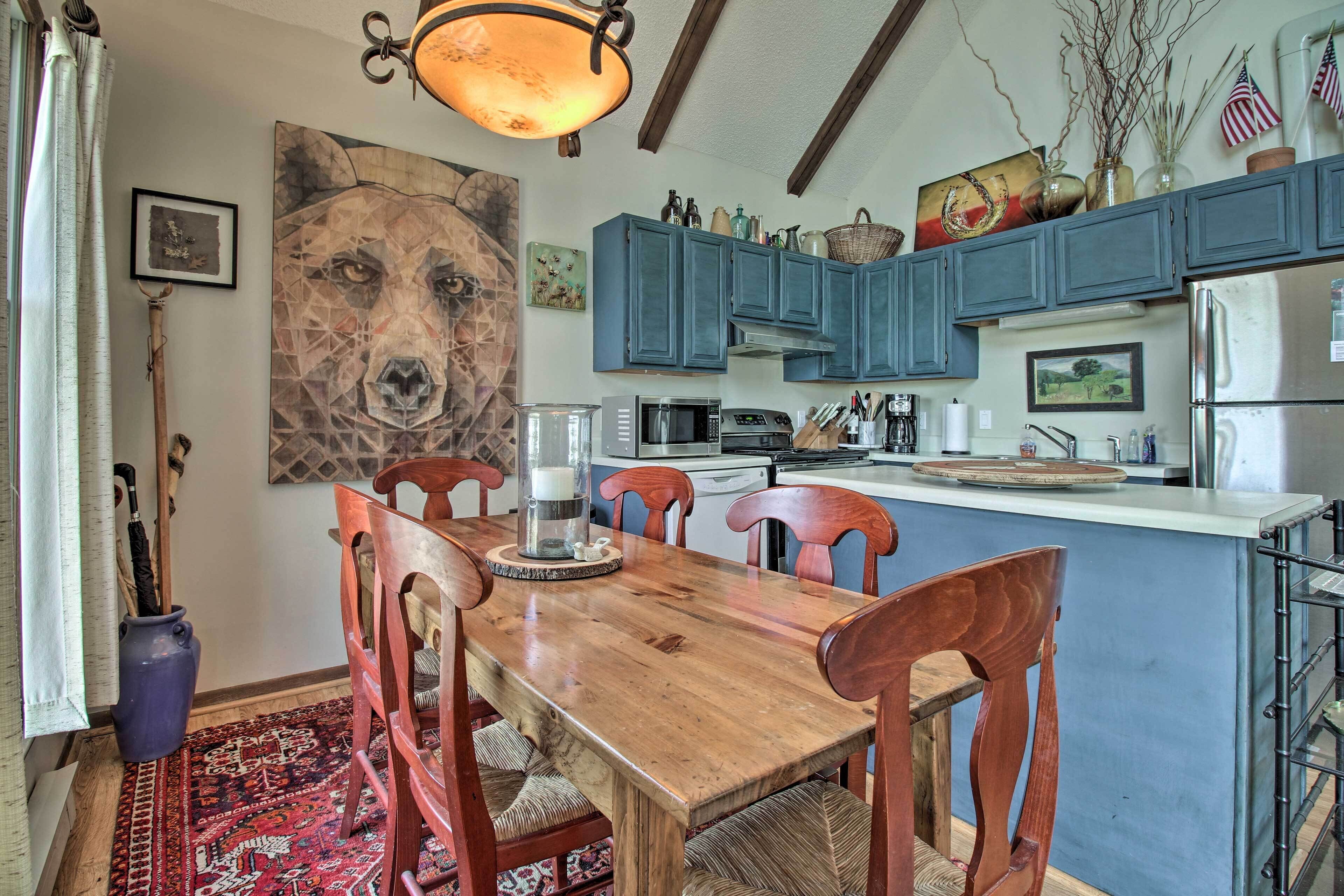 A dining area with a wooden table and chairs, a large abstract painting, and a kitchen with blue cabinets and various appliances.