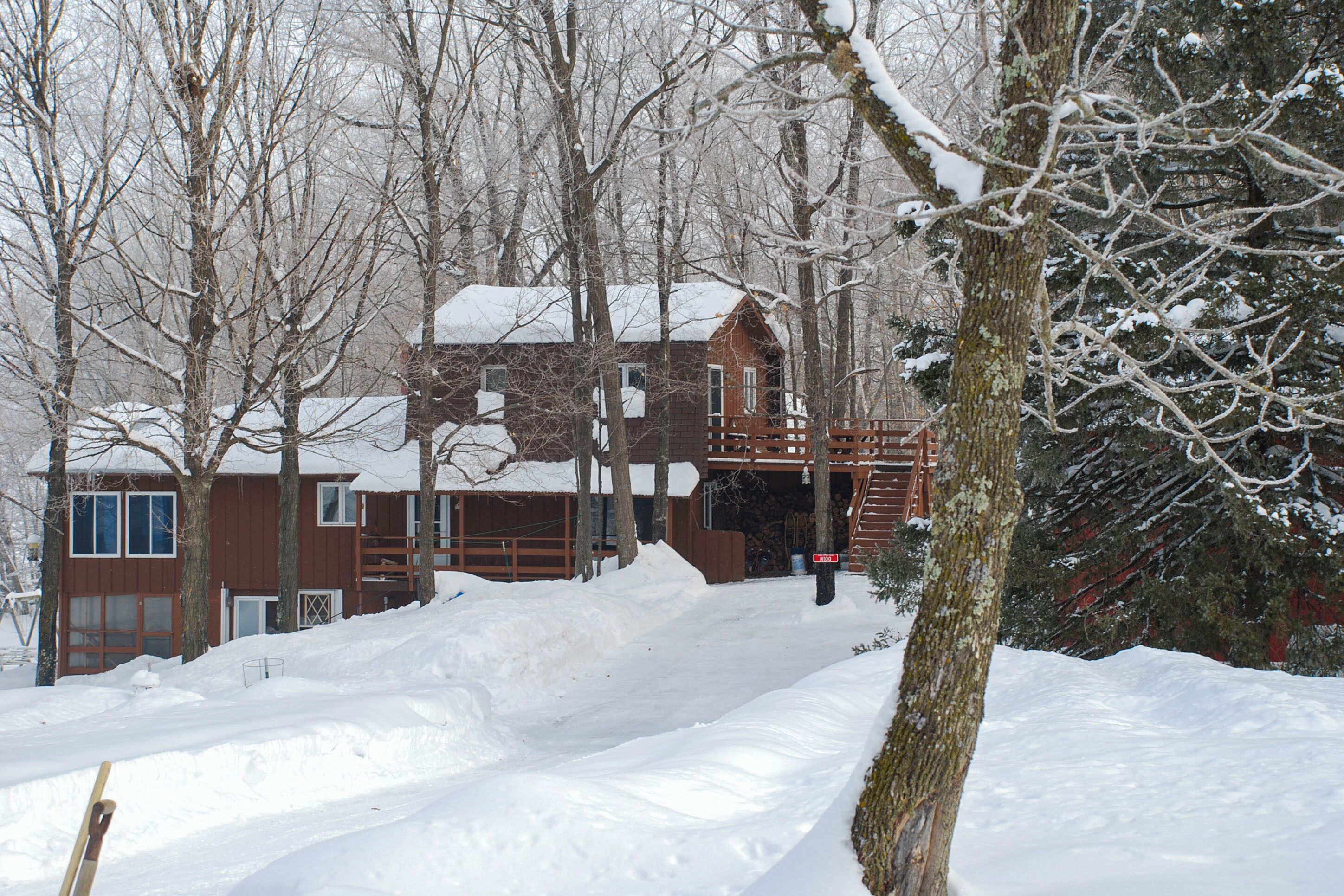 Eine Holzhütte mit einer hölzernen Terrasse und einem mit Schnee bedeckten Dach, umgeben von Bäumen und verschneitem Boden.