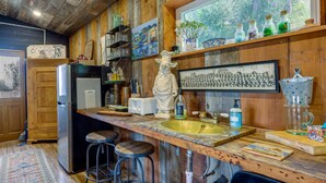 A rustic kitchen with wooden walls, a granite countertop, a refrigerator, a sink, bar stools, and various kitchen items.