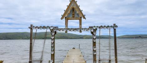 A wooden pier leading to a traditional structure with a pointed roof, surrounded by a body of water and hills in the background.