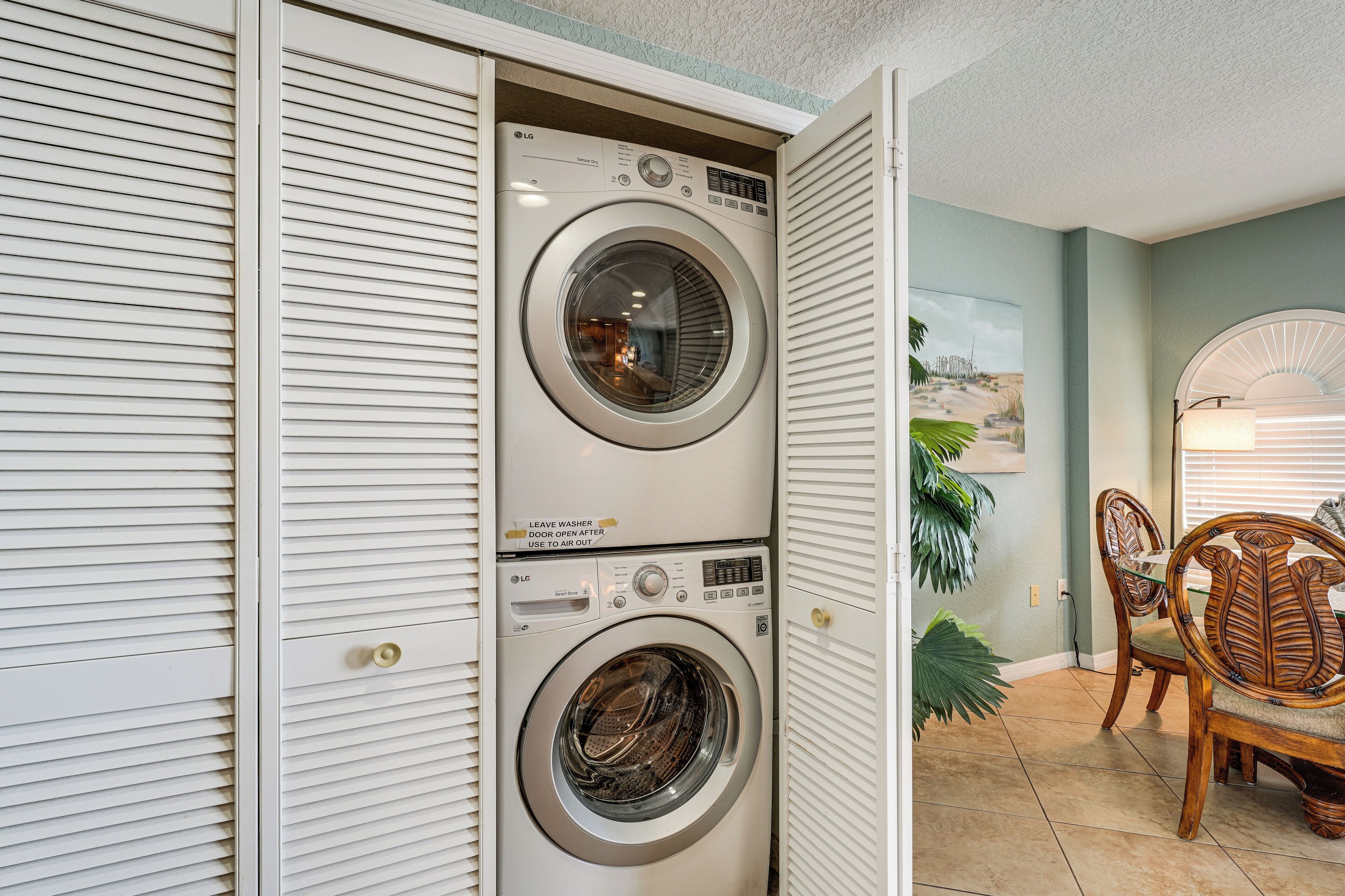 A laundry room with two stacked front-loading washers and dryers, a dining area with a wooden table and chairs, and a large potted plant.