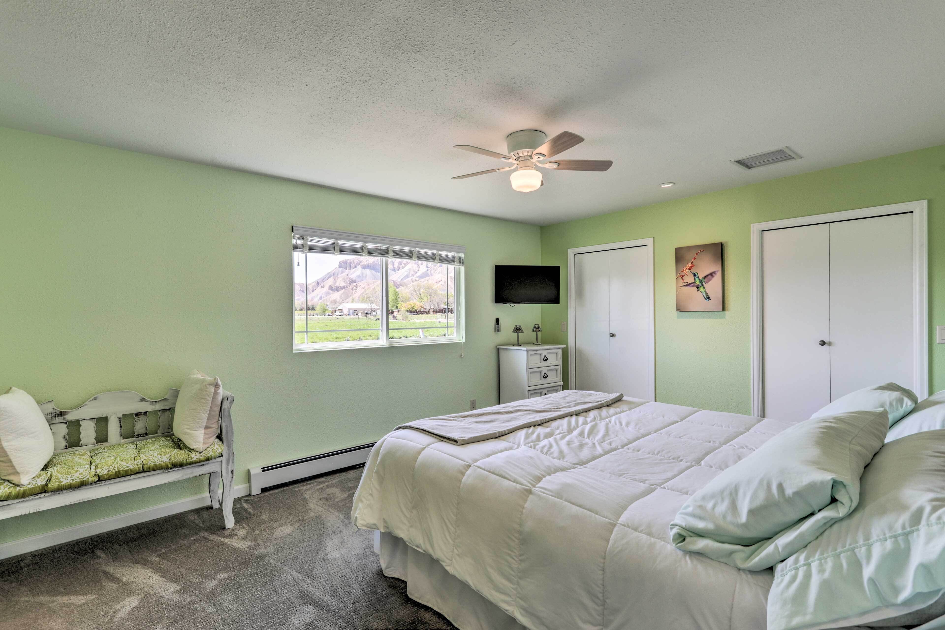 A bedroom with a bed, a bench, a ceiling fan, a window, a TV, and a door.