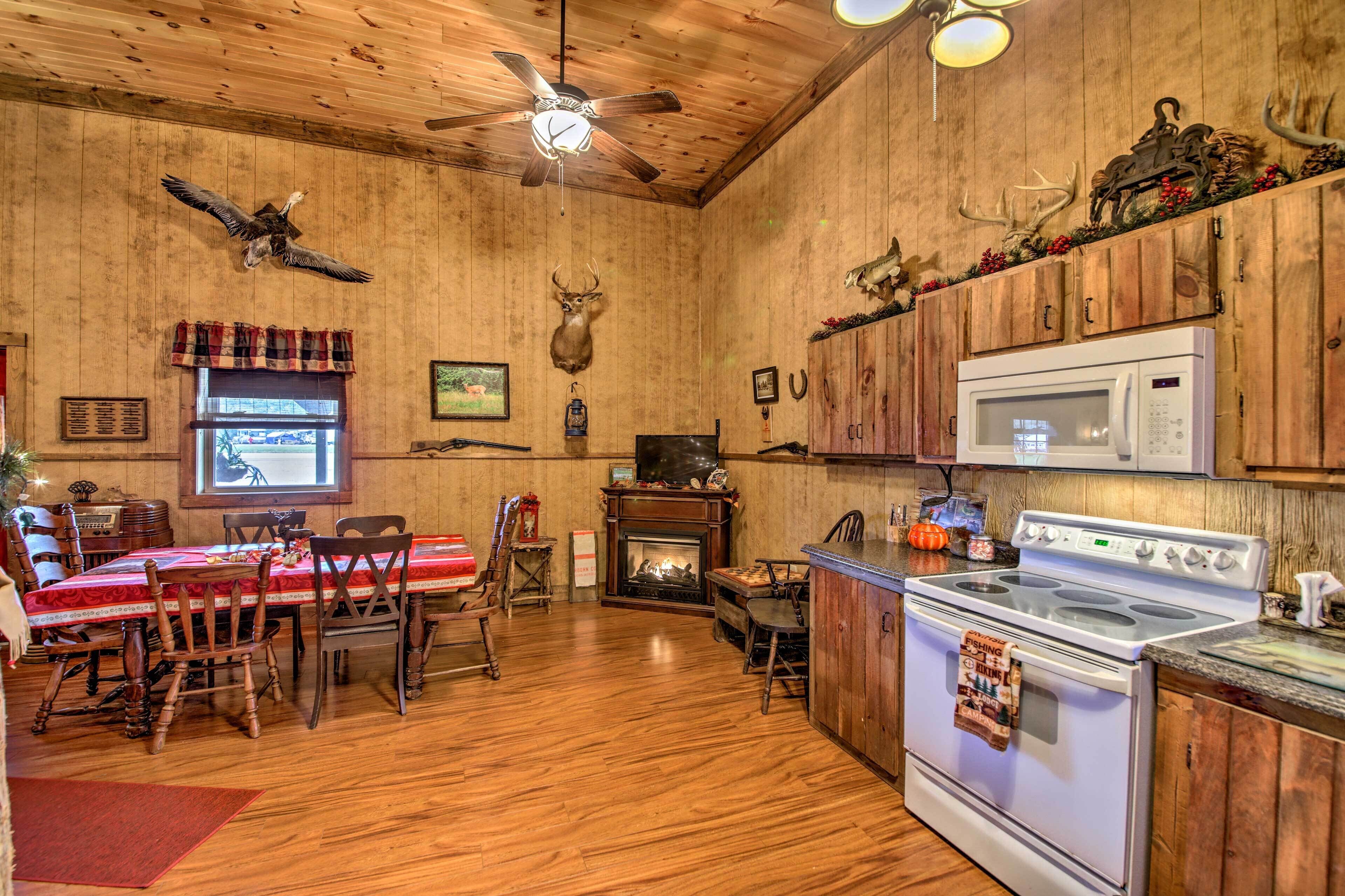 A rustic kitchen with wooden walls, a fireplace, a dining area with a red tablecloth, and a ceiling fan.