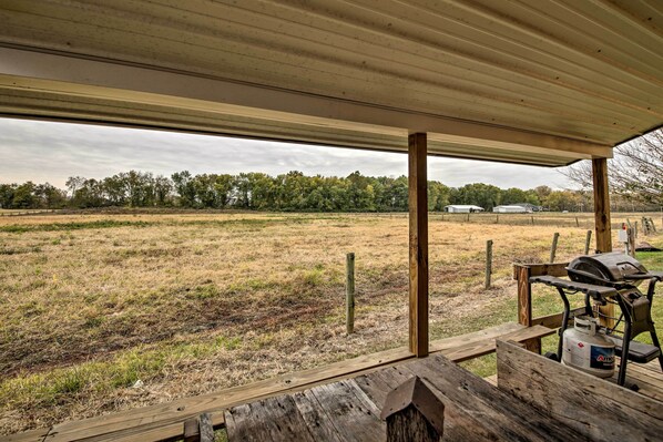 Un espacio al aire libre cubierto con una parrilla y tanque de propano, terraza de madera y vista a un campo y edificios lejanos.