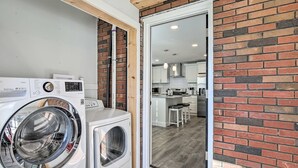 Een wasruimte met een wasmachine en droger, een bakstenen muur, en uitzicht op een moderne keuken met witte kastjes en barstoelen.