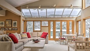 A spacious living room with a large sectional sofa, a coffee table, and a dining area with a table and chairs. There are large windows with a view of snow-covered trees. - Ski-in/ski-out Winter Park Condo w/ Pool Access! (Winter Park)