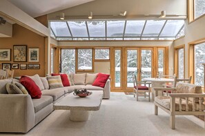 A spacious living room with a large sectional sofa, a coffee table, and a dining area with a table and chairs. There are large windows with a view of snow-covered trees. - Ski-in/ski-out Winter Park Condo w/ Pool Access! (Winter Park)