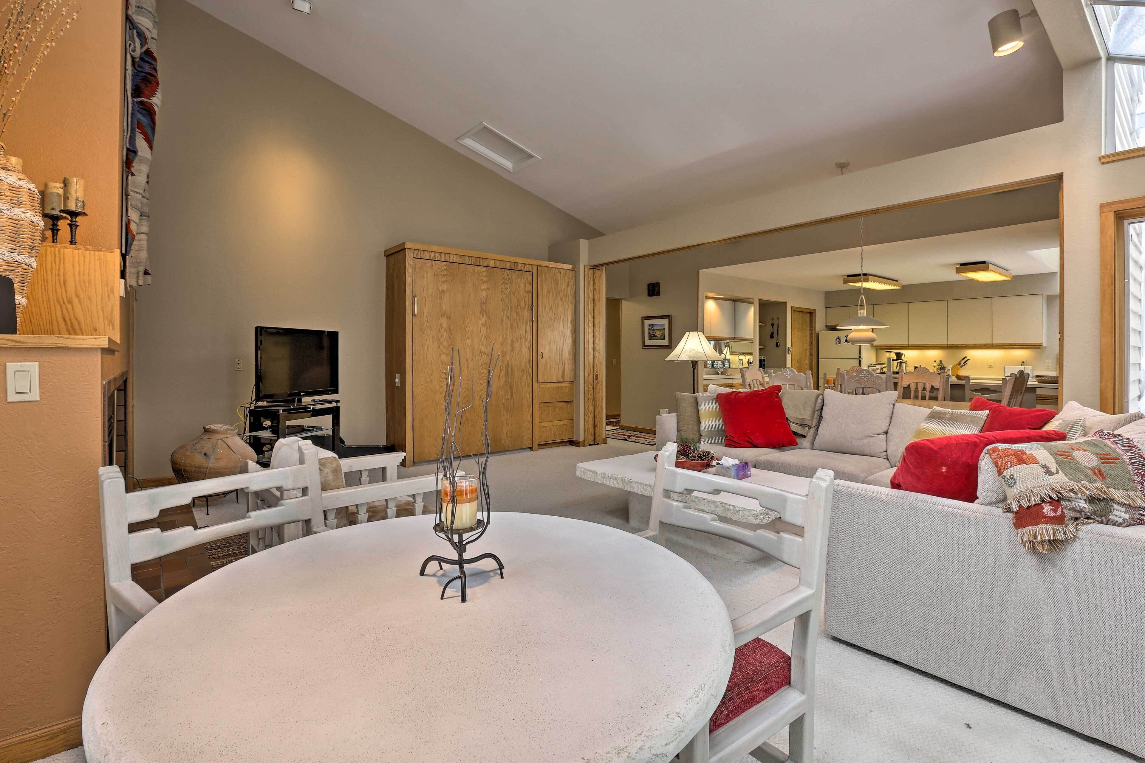 A living room with a round table, white chairs, a sofa with red cushions, and a television.