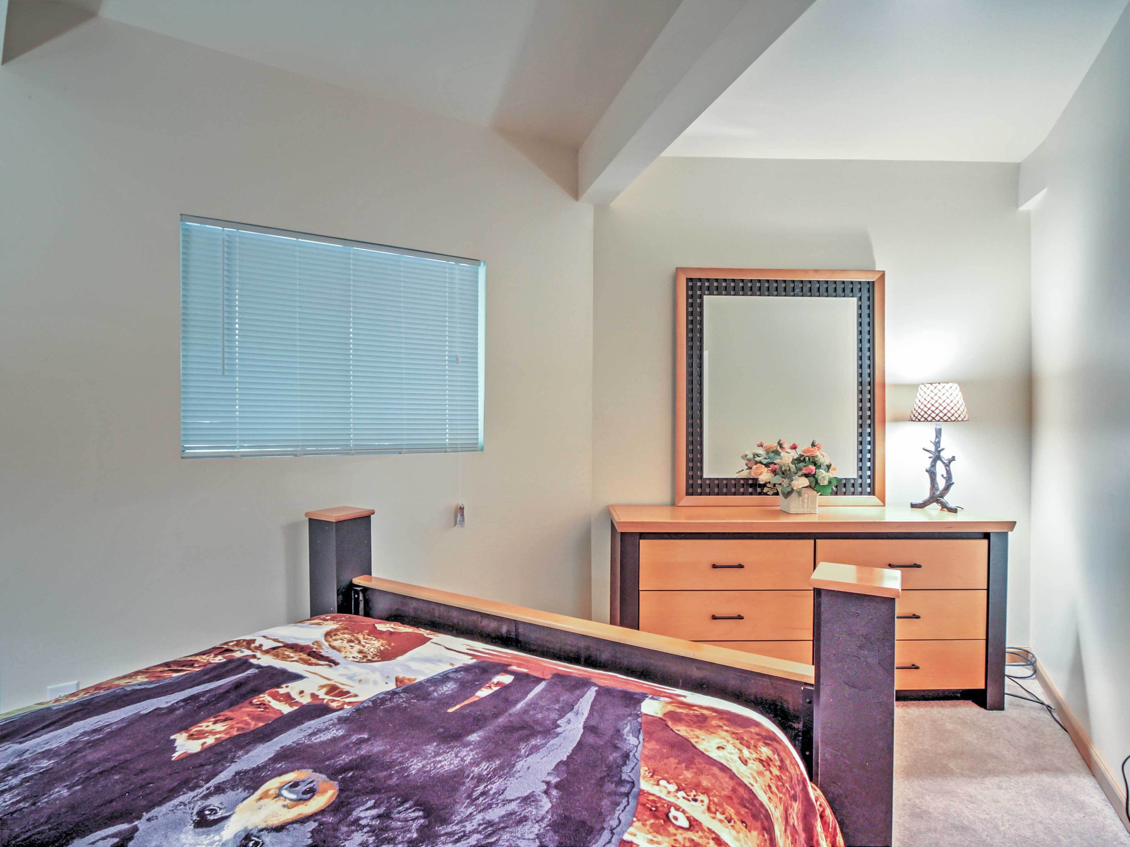 Ein Schlafzimmer mit Bett, Kommode, Spiegel, Lampe und Fenster mit Jalousien.