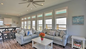 A spacious living room with a large window, a ceiling fan, and a dining area with a kitchen in the background. - Crystal Tides - Stunning Home W/oceanfront Views (Port Bolivar)