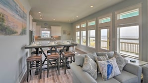 A spacious living area with a gray couch, a dining table with stools, and large windows offering a view of the ocean. - Crystal Tides - Stunning Home W/oceanfront Views (Port Bolivar)
