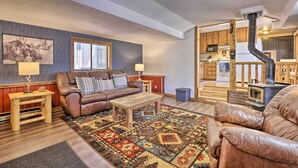 A cozy living room with a brown leather sofa, a wooden coffee table, and a patterned rug. There is a fireplace with a chimney, wooden cabinets, and a window with blinds. - Fireplace, Deck & Mtn Views: Grand Lake Home! (Grand Lake)