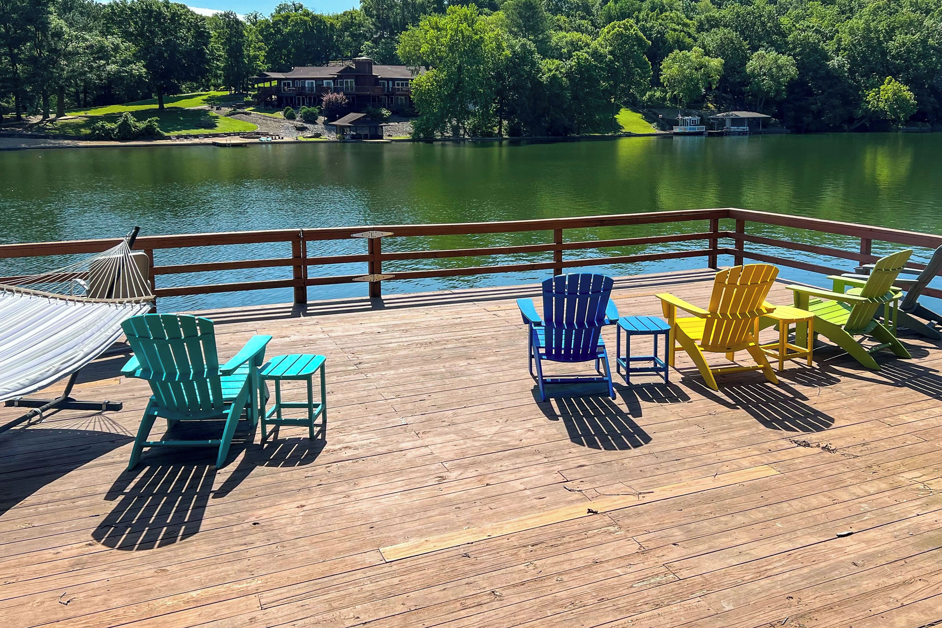 A wooden deck with colorful Adirondack chairs and a hammock overlooking a lake.