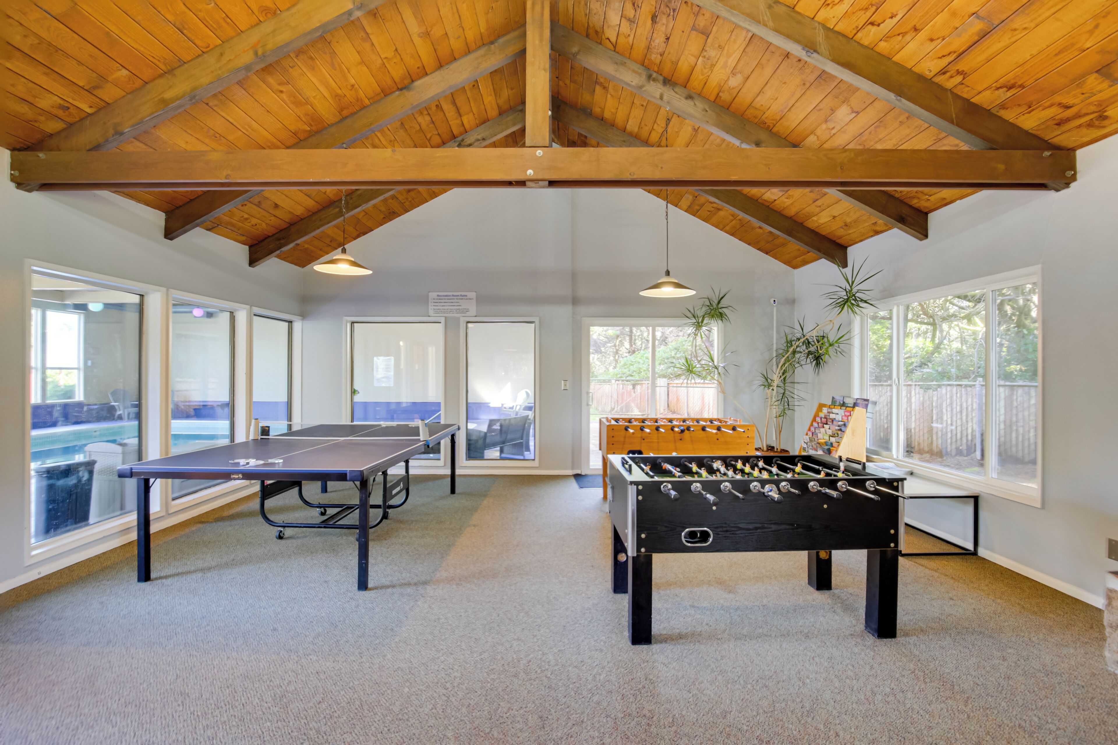 A spacious indoor area with a ping pong table, a foosball table, and large windows offering views of a backyard.