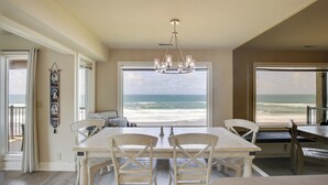 A dining area with a view of the ocean, featuring a white table and chairs, a chandelier, and a large window. - Oceanfront Oregon Retreat - Pool, Sauna & Patio! (Lincoln City)