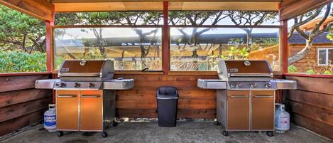 A covered outdoor area with two barbecue grills, a trash bin, and propane tanks.