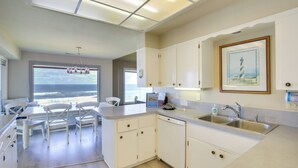 A modern kitchen with white cabinetry, a large window overlooking the ocean, and a dining area with white chairs. - Oceanfront Oregon Retreat - Pool, Sauna & Patio! (Lincoln City)
