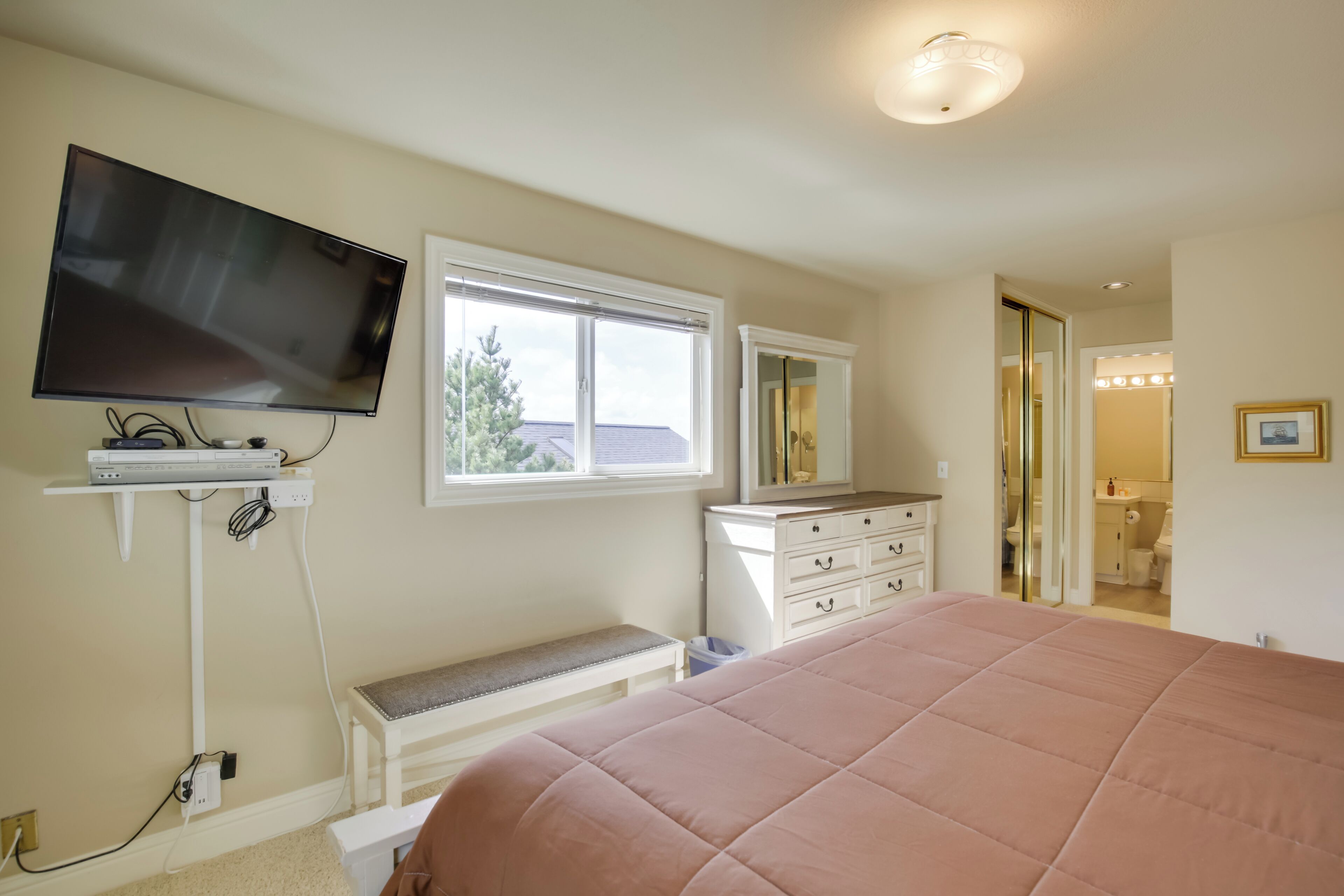 Ein Schlafzimmer mit einem großen Bett, einem an der Wand montierten Fernseher, einem Fenster, einem Schminktisch mit Spiegel und einer Tür zu einem Badezimmer.