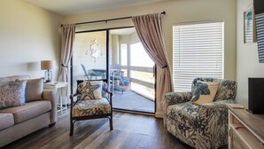 Ein Wohnzimmer mit einer Couch, zwei gemusterten Sessel, einem Couchtisch und einem Fernseher. Eine Glasschiebetür führt zu einem Außenbereich mit Tisch und Stühlen.