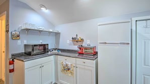 Cucina con pensili bianchi, forno a microonde, tostapane e frigorifero. Sopra il bancone, mensole con bicchieri e un piccolo oggetto decorativo.