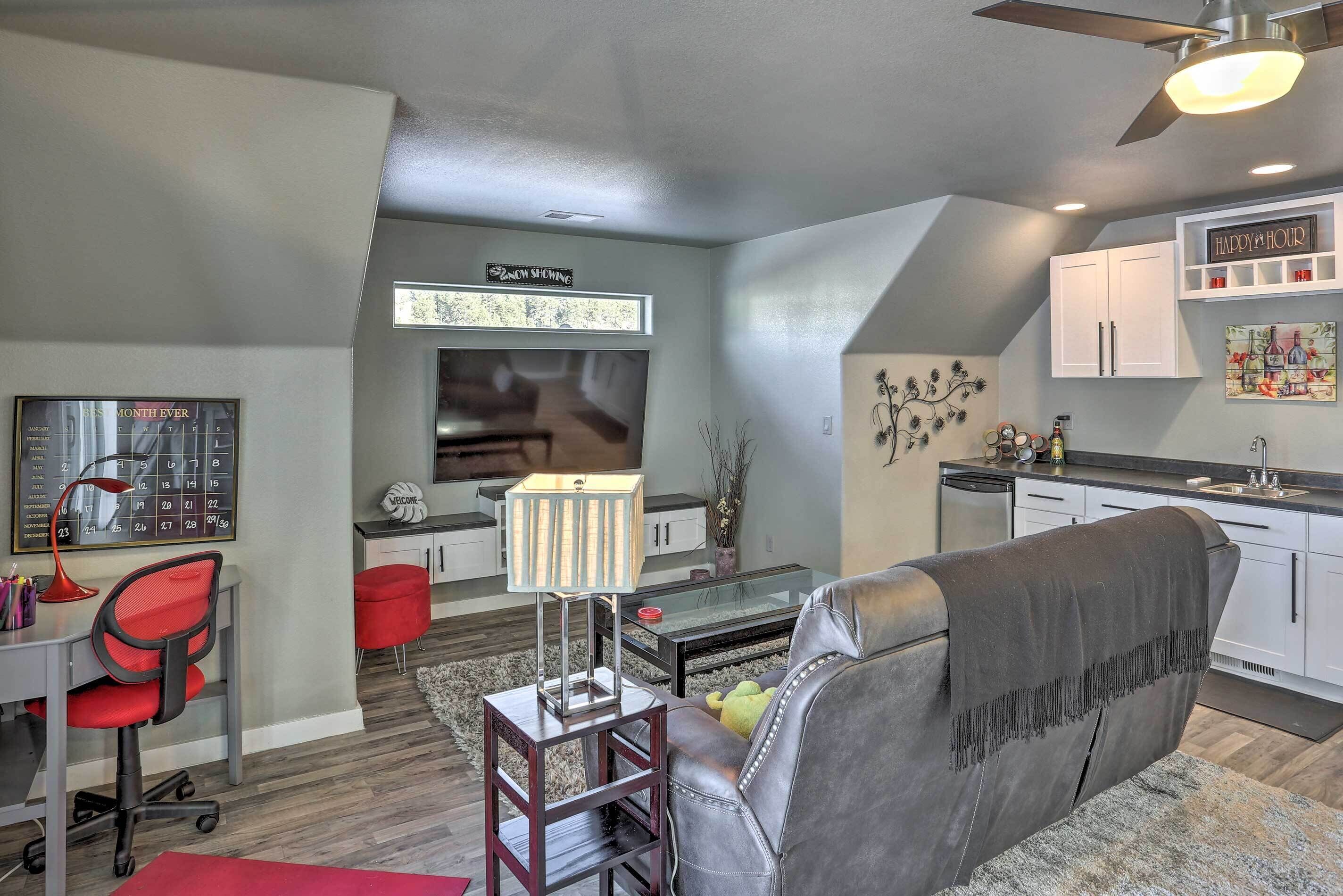 A modern living room with a leather sofa, a desk with a red chair, a glass coffee table, and a lamp. There is a kitchen area with white cabinets and a ceiling fan.