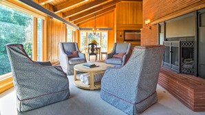 A cozy room with wooden ceiling beams, a fireplace, and large windows offering a view of the outdoors. - Hot Tub & Sauna! 7-acre Coastal Michigan Home (Ludington)