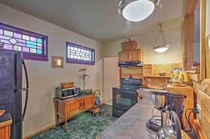 A compact kitchen with a refrigerator, microwave, stove, and coffee maker. There are wooden cabinets and a window with stained glass. - Bozeman Cottage w/ Mountain Views < 6 Mi to Dtwn! (Bozeman)