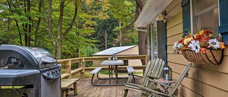 Une terrasse en bois équipée d’un barbecue, d’une table de pique-nique et de chaises, le tout entouré d’arbres.