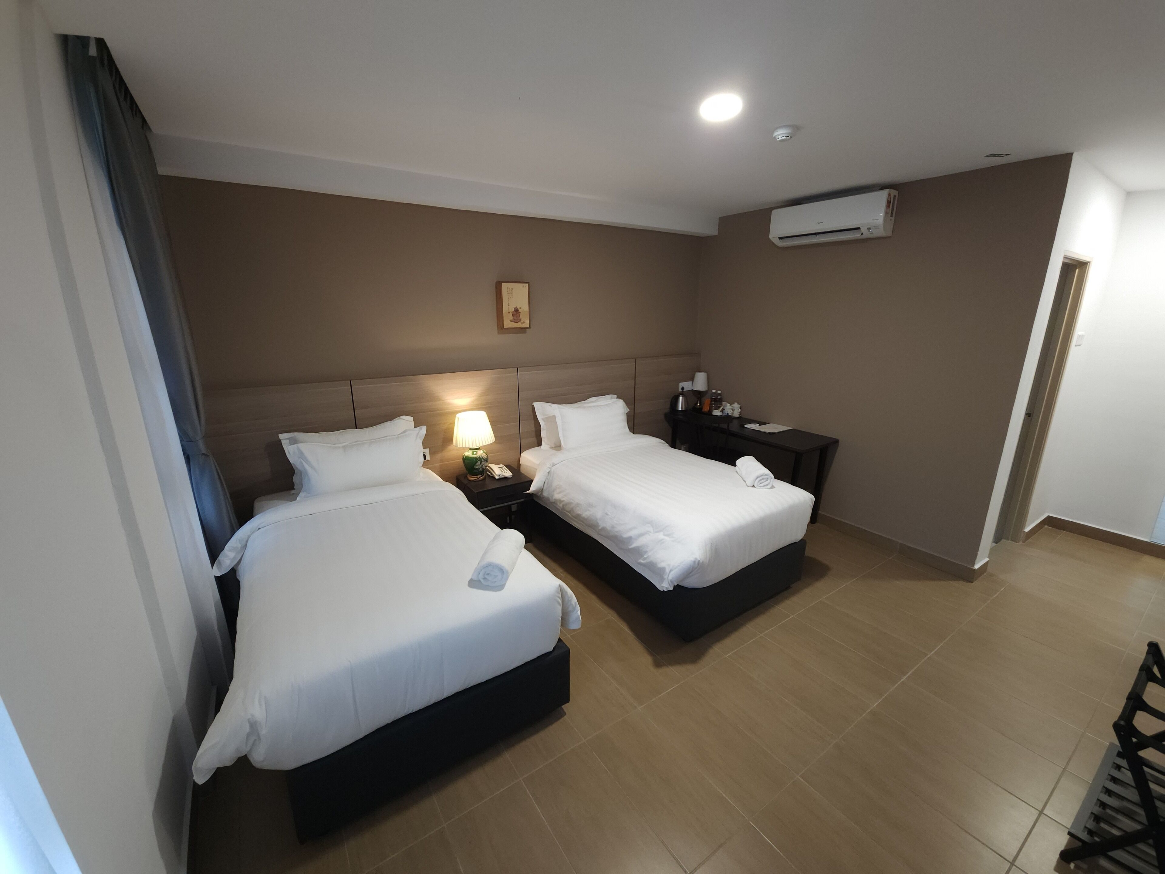 A hotel room with two beds, a desk, a chair, a nightstand, a lamp, a picture on the wall, and an air conditioning unit.