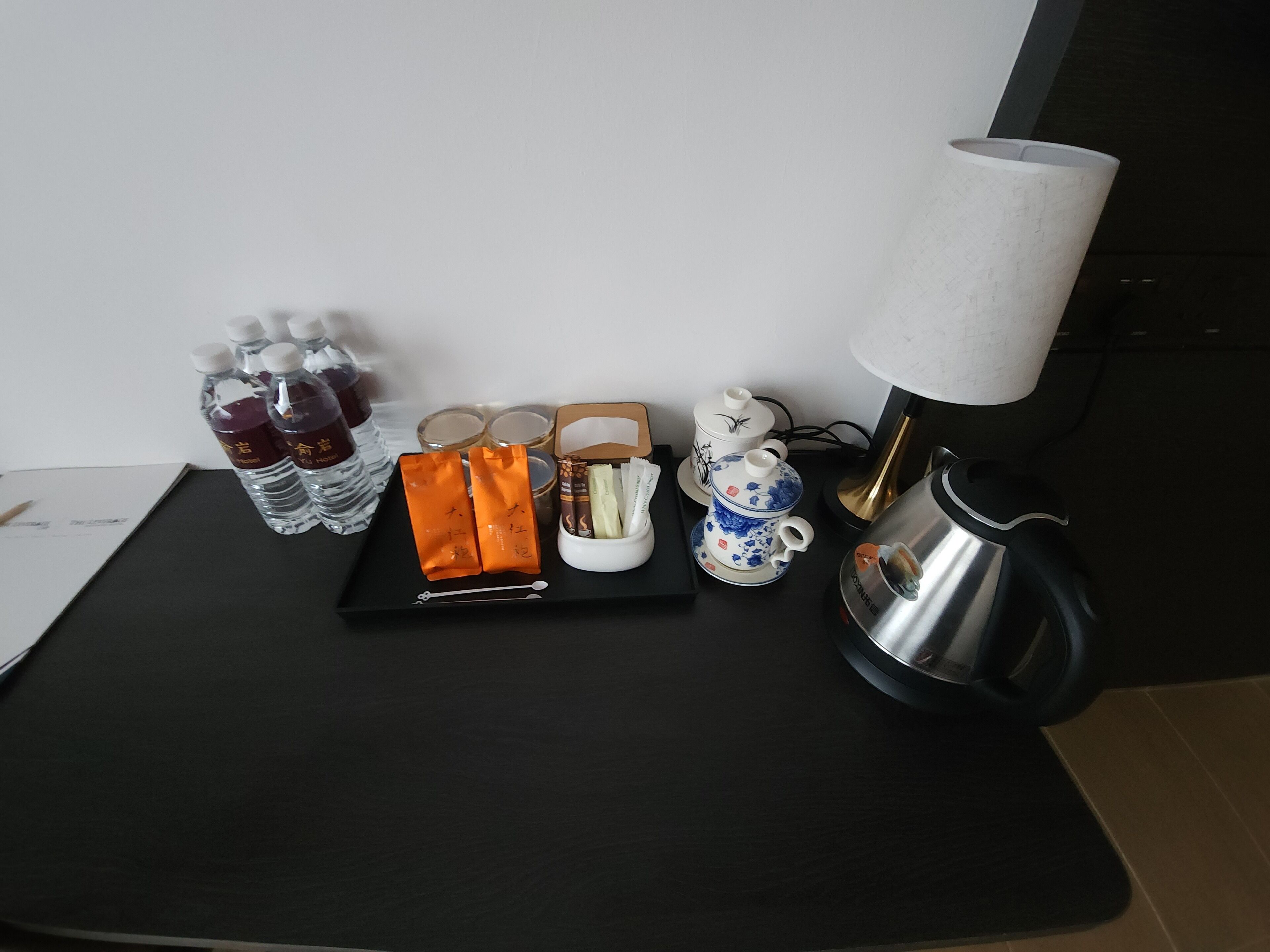 Een tafel met een zwarte dienblad waarop waterflessen, oranje zakjes, een theepot, een kopje en een waterkoker staan.