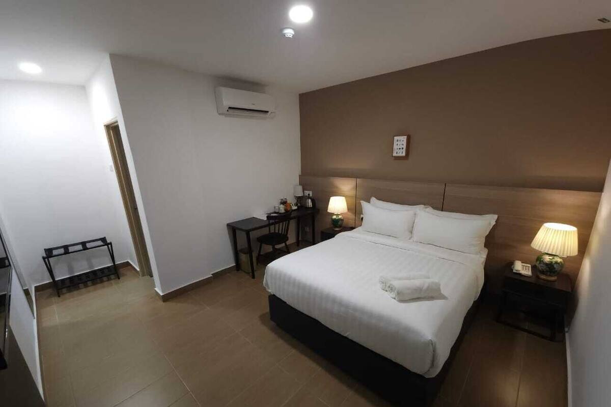 A hotel room with a large bed, bedside tables, a desk, and an air conditioning unit.