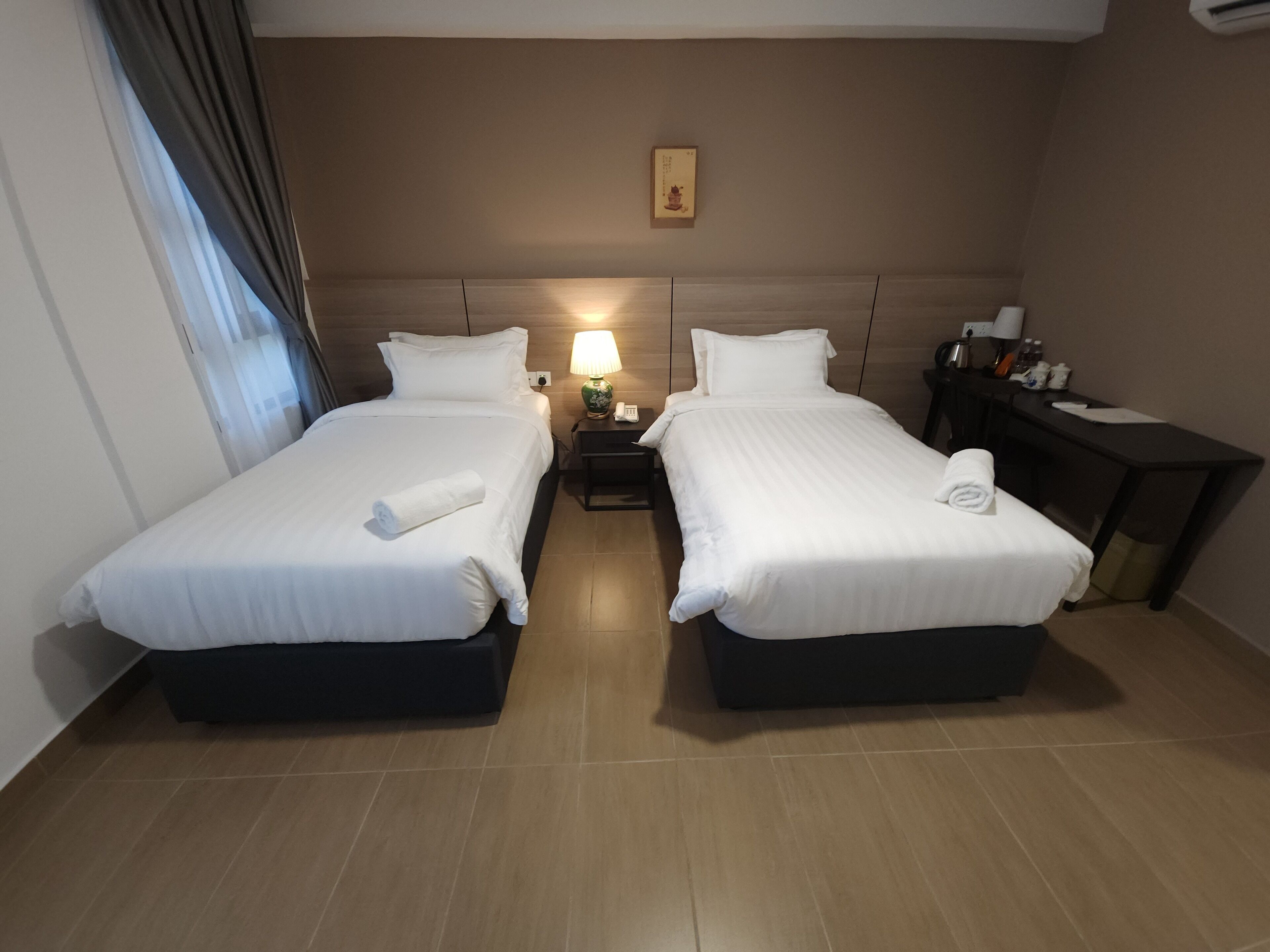 A hotel room with two beds, a nightstand with a lamp, a desk, and a small picture on the wall.
