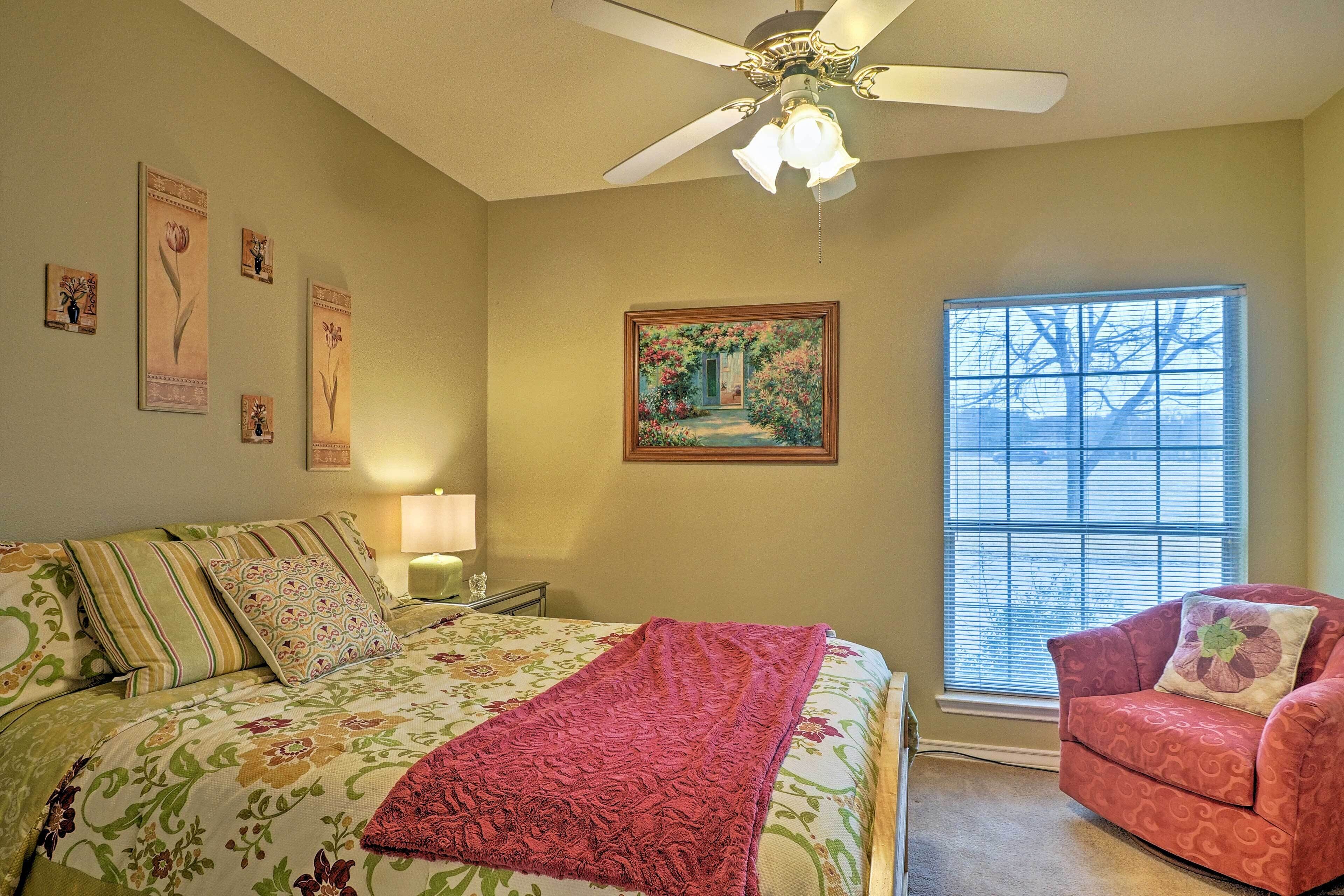 A bedroom with a bed, a chair, a window with blinds, a ceiling fan, and wall art.