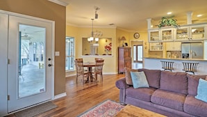 A spacious living room with a wooden floor, a purple sofa with cushions, a dining area with a wooden table and chairs, and a kitchen with white cabinets and a plant on the counter. - Round Rock Lakefront Home w/ Water Toys & Fire Pit (Round Rock)