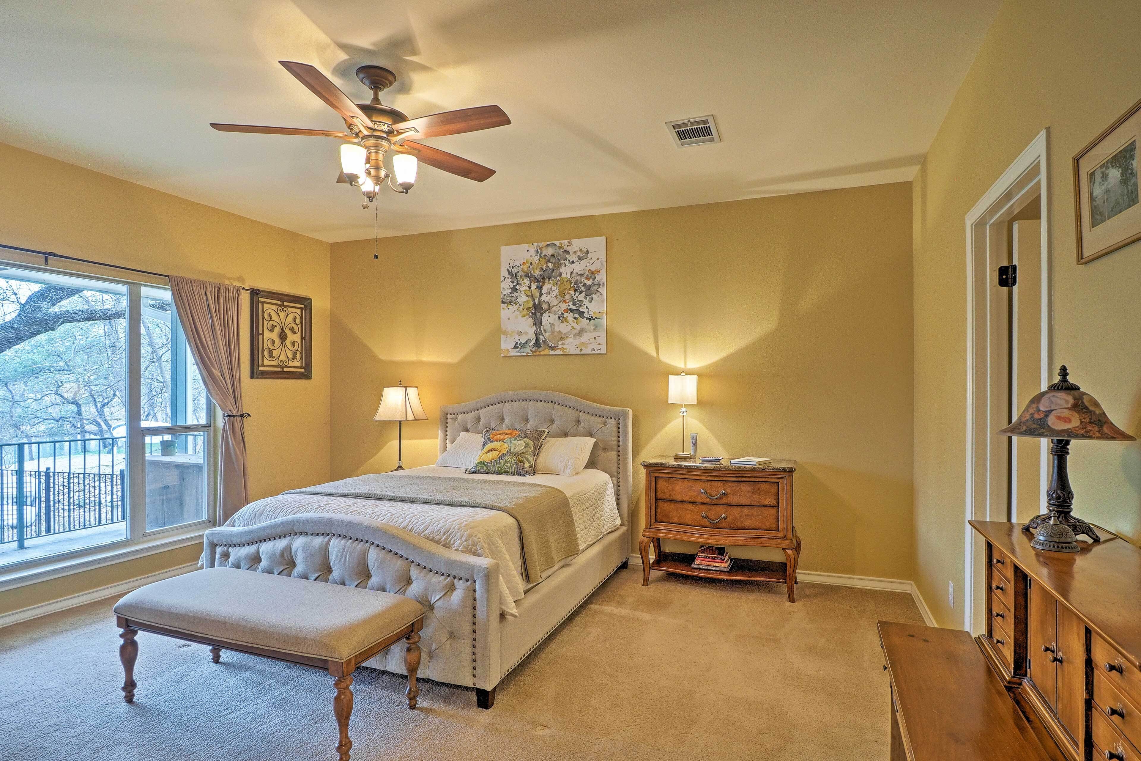 A bedroom with a large bed, a ceiling fan, a window with curtains, a bedside table with lamps, and a dresser.