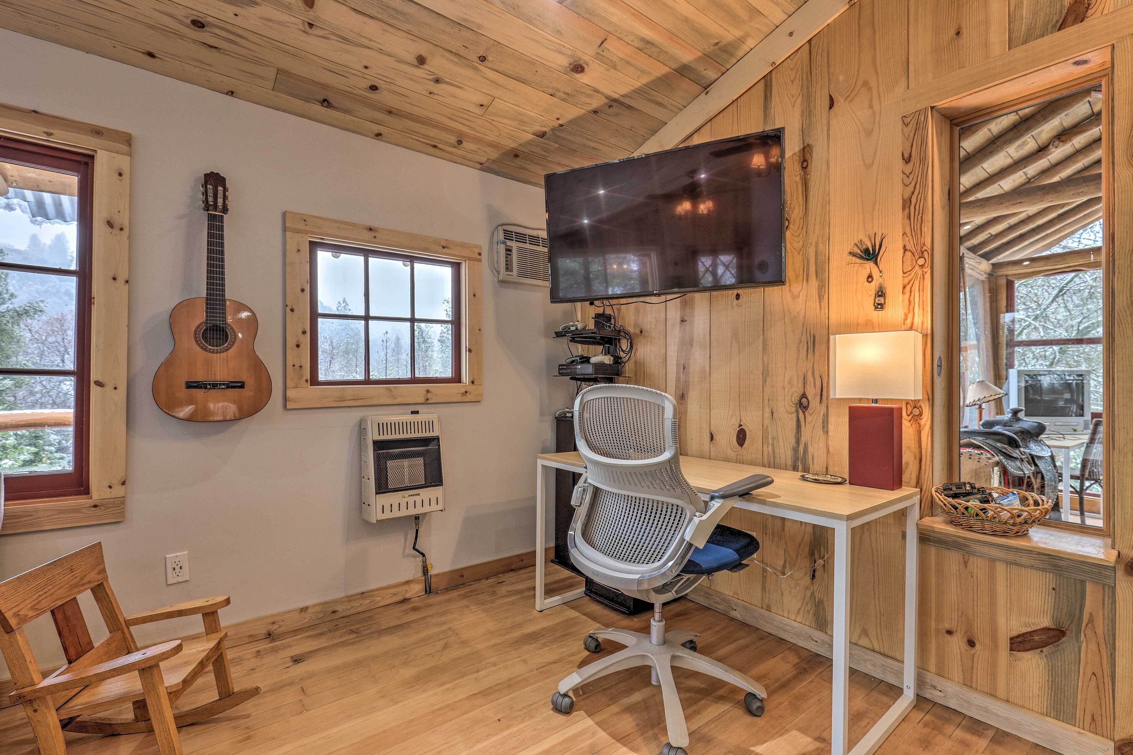 A room with wooden walls, a desk with a chair, a mounted TV, a guitar, and windows with a view of trees.