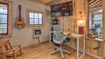 A room with wooden walls, a desk with a chair, a mounted TV, a guitar, and windows with a view of trees.