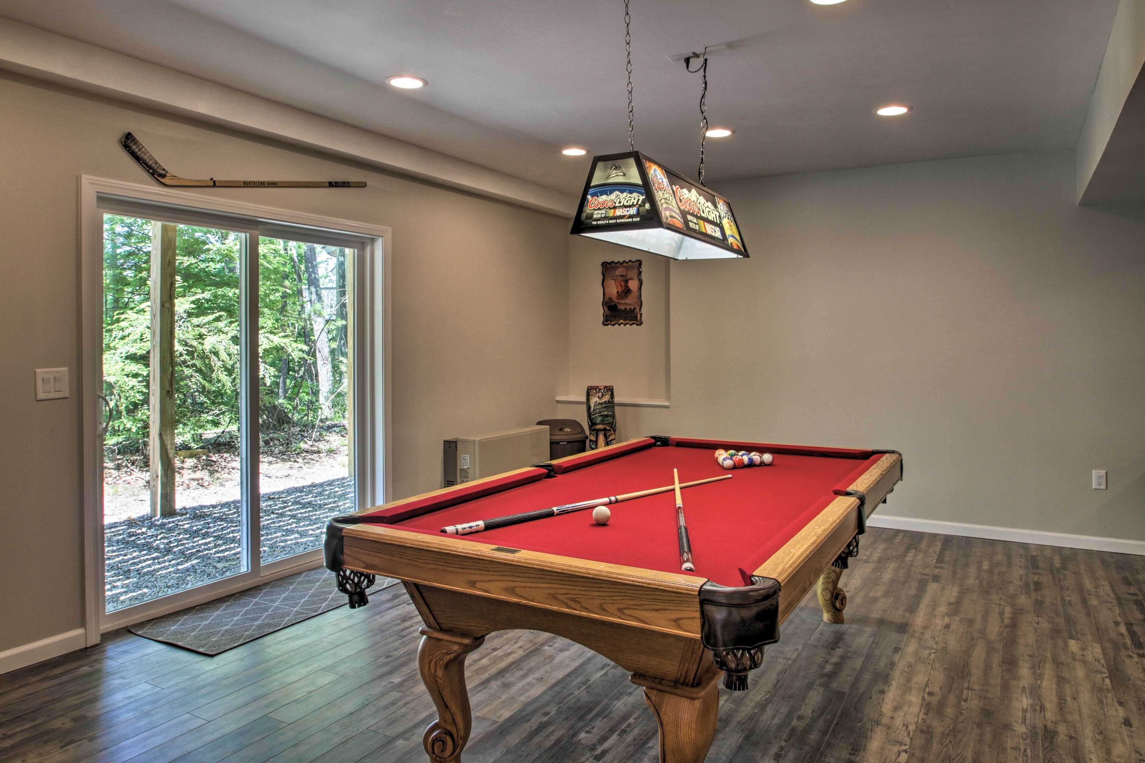A pool table in a room with wooden flooring, a ceiling fan, and a view of trees through a sliding glass door.