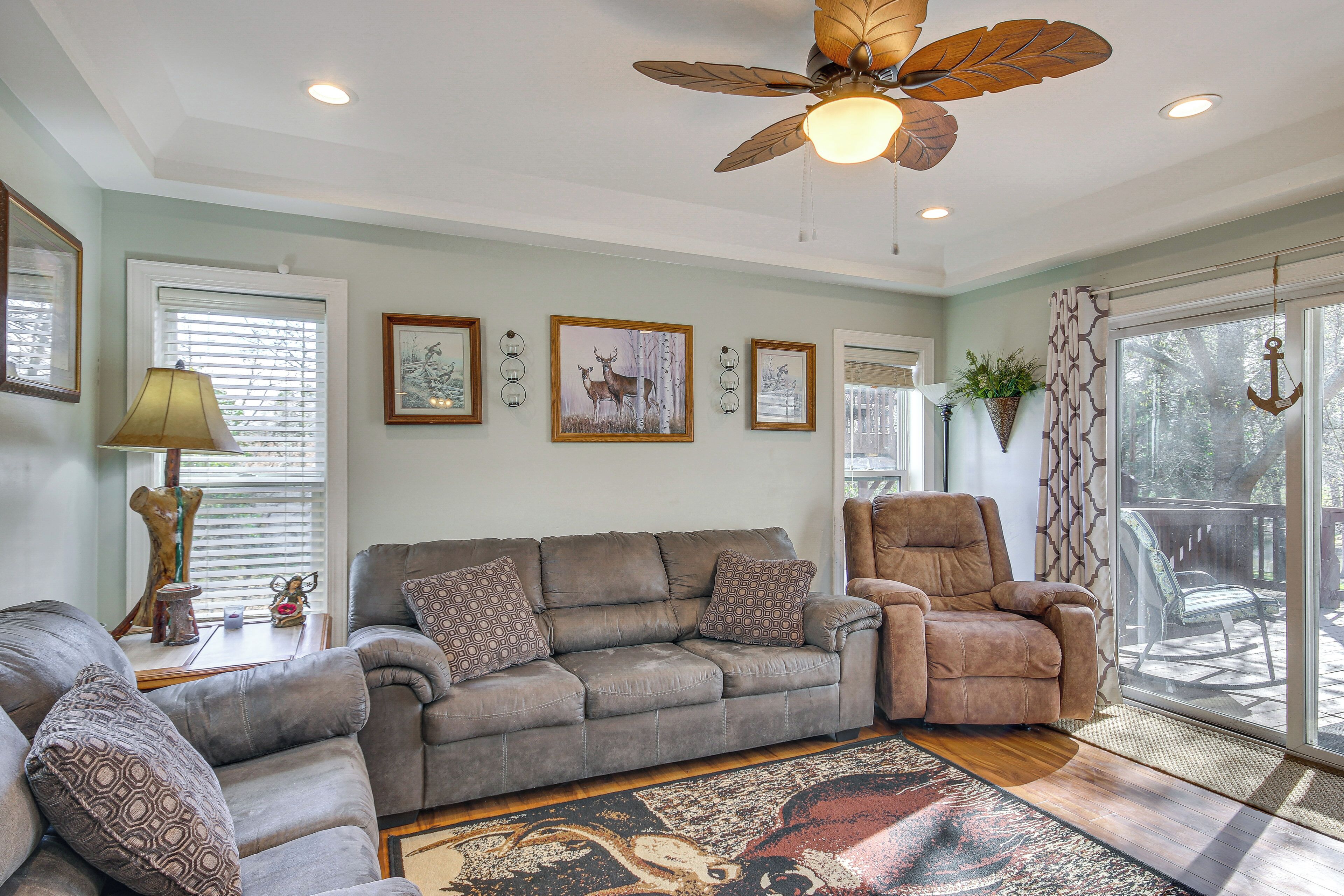 A living room with a sofa, armchair, and a ceiling fan. There are framed pictures on the wall and a window with blinds.