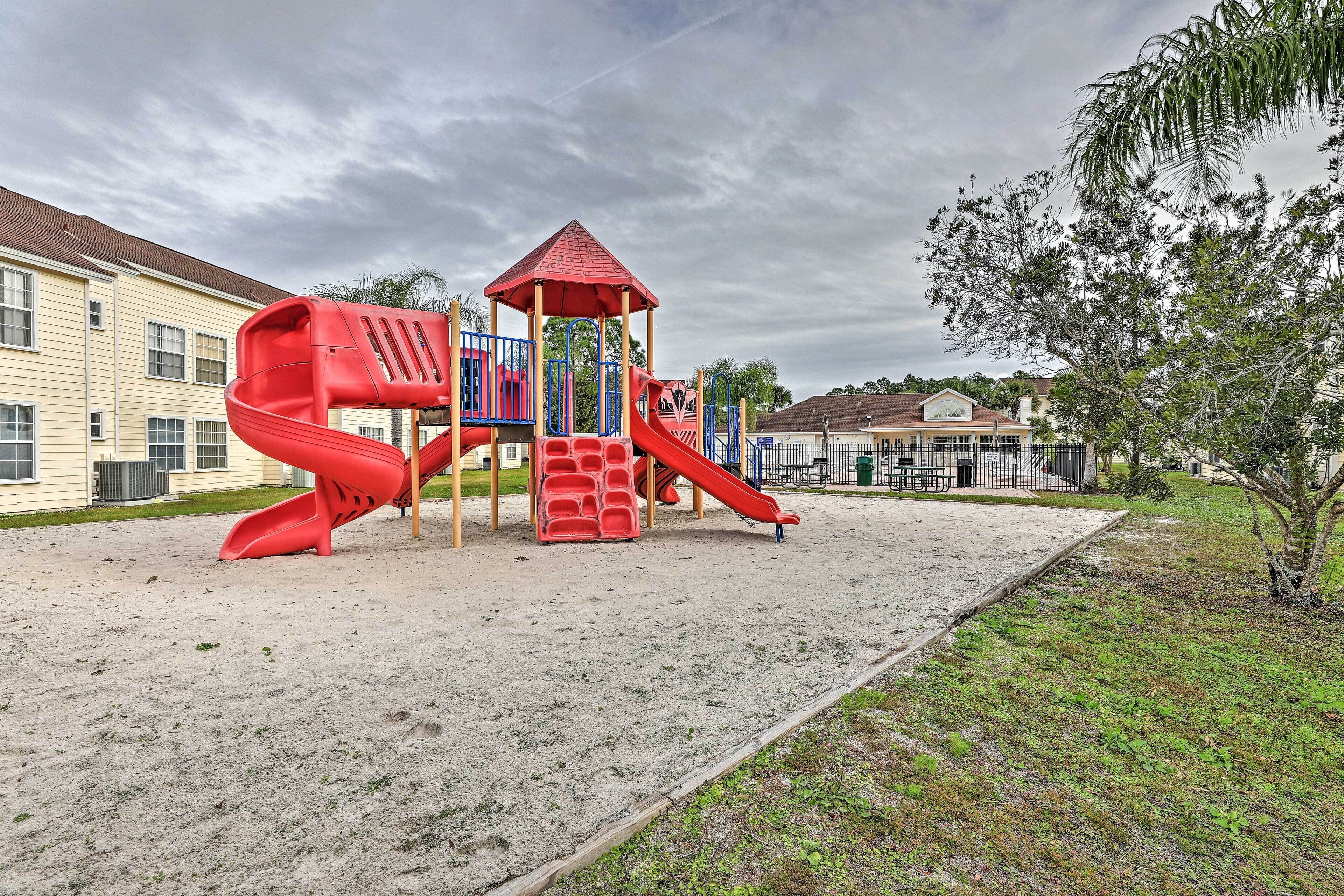 Un parque infantil con un tobogán rojo y estructuras para escalar, rodeado de arena y edificios residenciales.