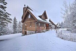 A wooden cabin with a steep roof, surrounded by snow-covered trees and a snow-covered driveway.