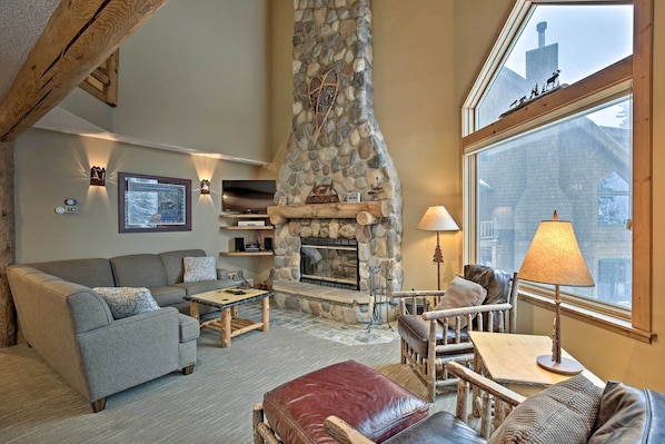 A cozy living room with a stone fireplace, a sofa, a leather ottoman, and a wooden coffee table.