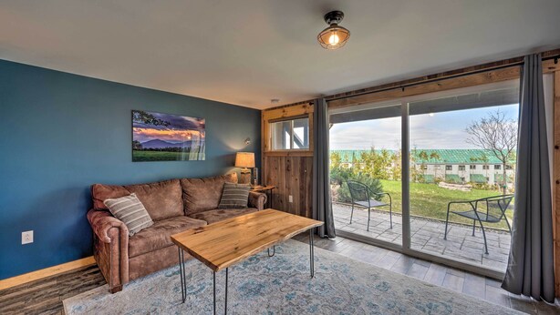 A living room with a brown leather sofa, a wooden coffee table, and a large sliding glass door leading to an outdoor patio with a view of a building and greenery. - Slopeside Burke Mountain Condo w/ Fireplace! (East Burke)