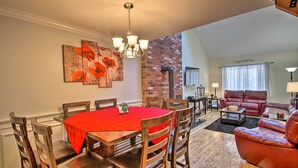 A dining area with a red tablecloth, wooden chairs, and a chandelier. There is a brick wall and a living area with a leather sofa and a fireplace. - Penthouse Ski Unit ~ 1 Mi to Attitash Mountain! (Bartlett)