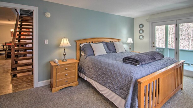 A bedroom with a bed, nightstands, a lamp, a wooden dresser, a staircase, and a view of trees through a glass door. - Penthouse Ski Unit ~ 1 Mi to Attitash Mountain! (Bartlett)