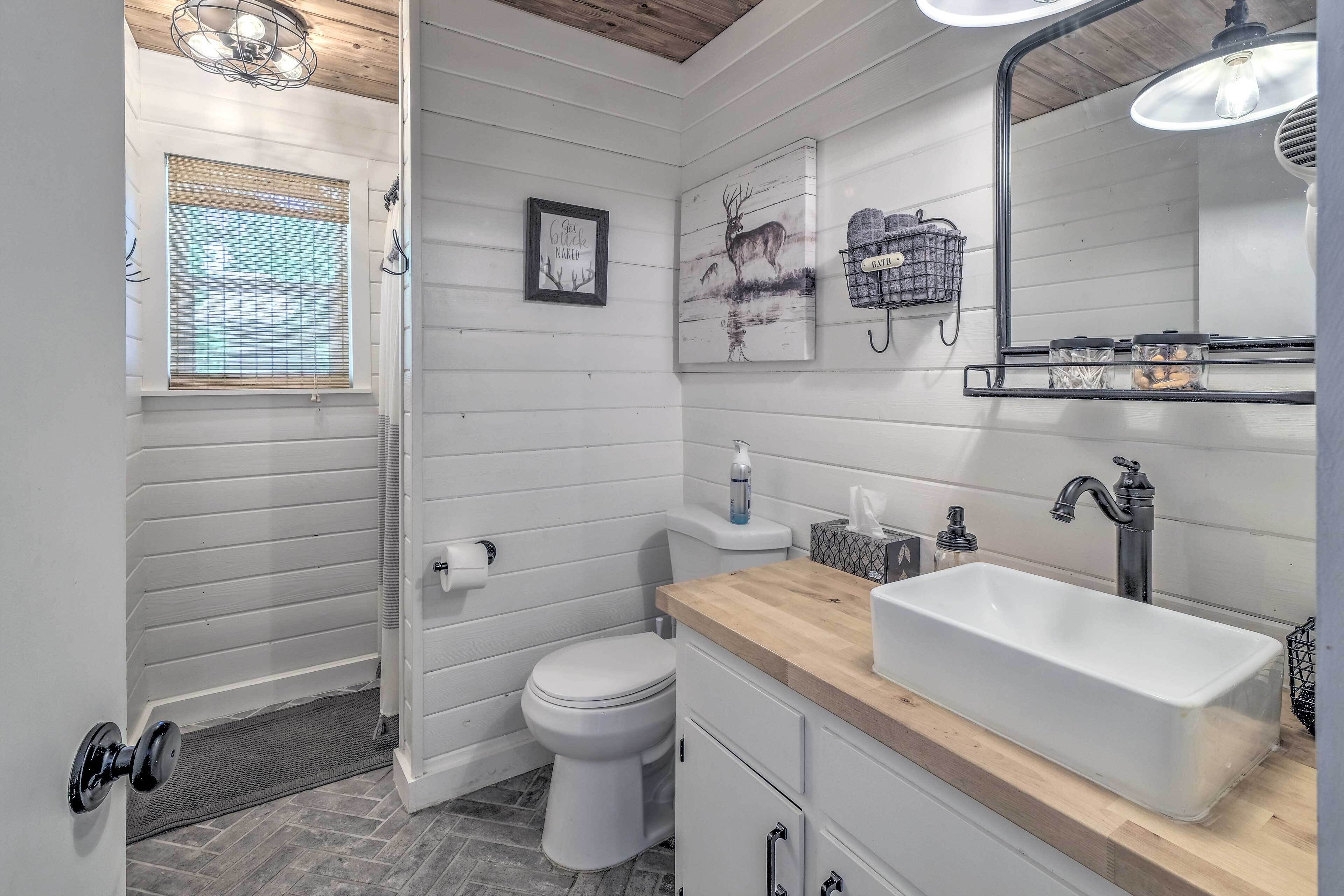 Une salle de bain avec un lavabo, des toilettes et une douche, le tout de couleur blanche. On y trouve une fenêtre avec des stores, un tableau encadré au mur et un miroir au-dessus du lavabo.