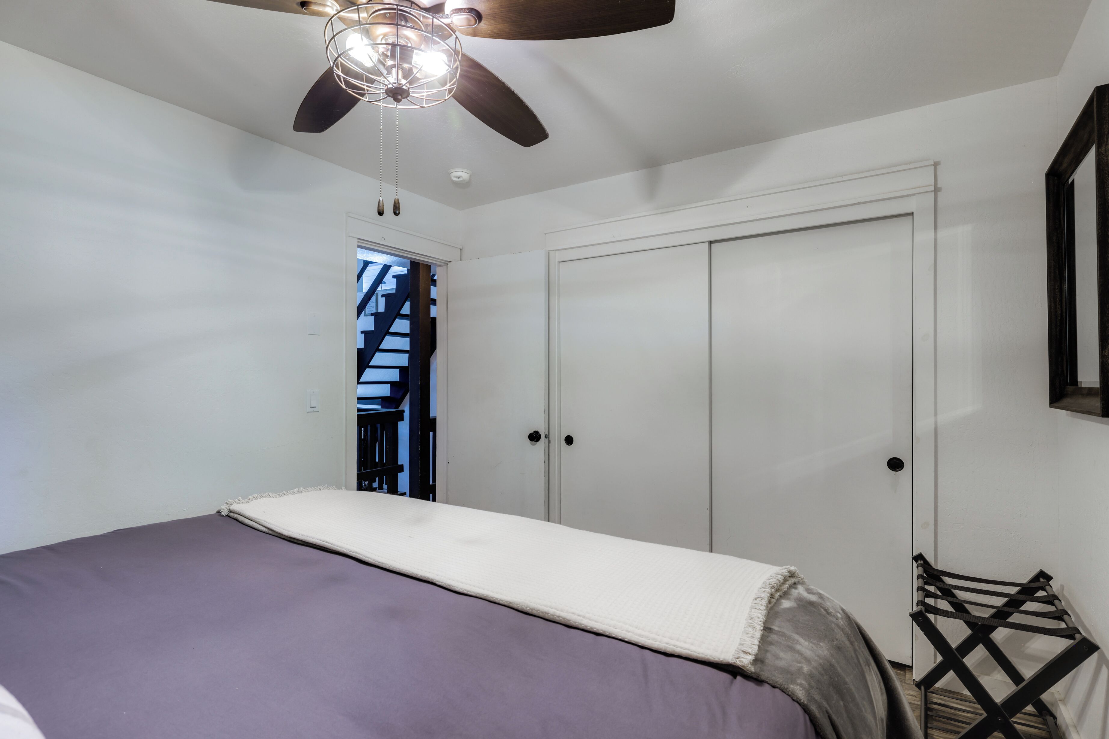 A bedroom with a bed, a ceiling fan, a wardrobe, and a view of a staircase.
